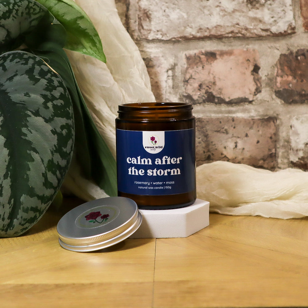 Candle jar labeled 'calm after the storm' on a wooden surface with a plant and fabric in the background.