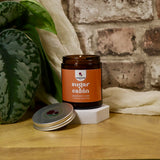 Jar of 'sugar cabin' candle with a plant and fabric in the background