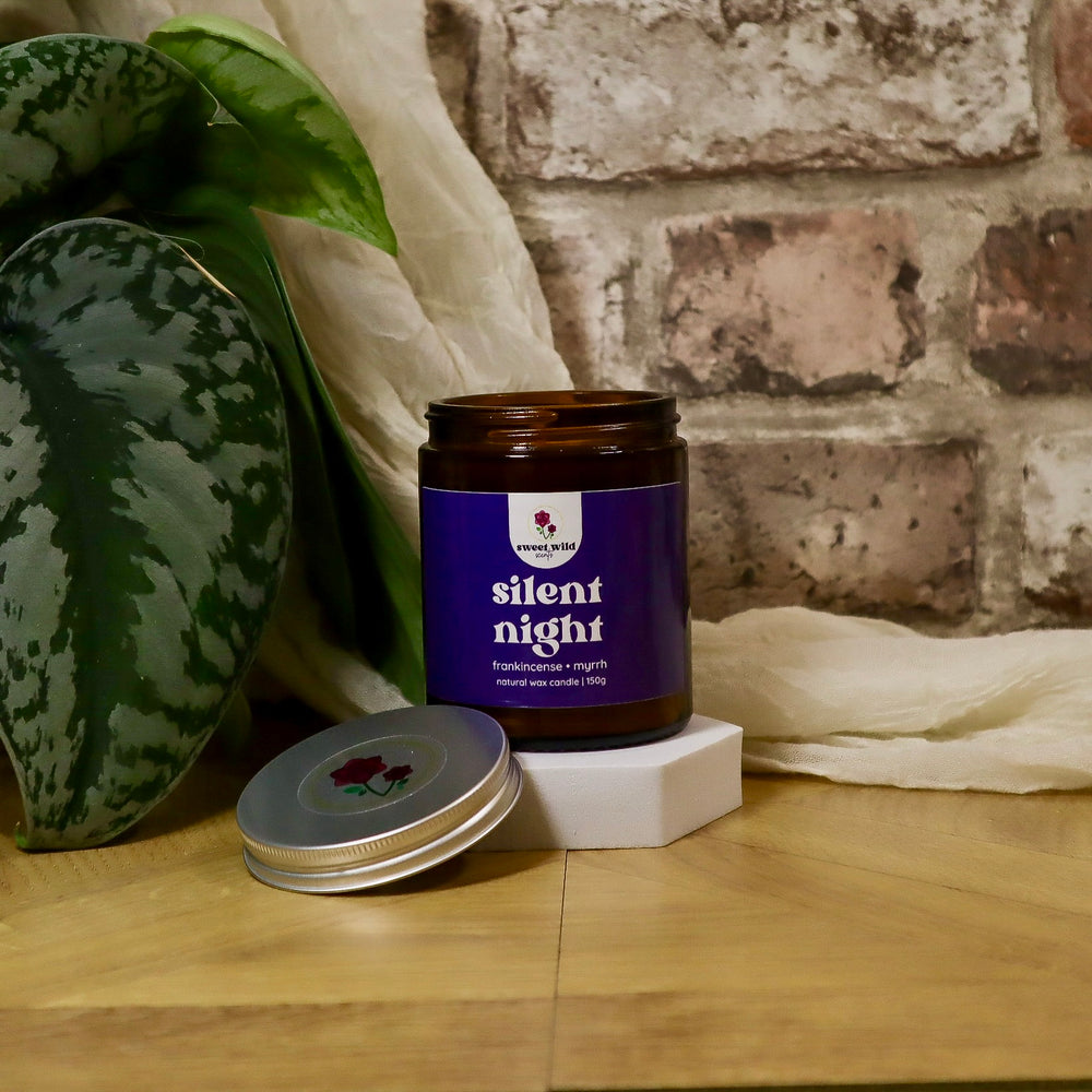 Candle jar labeled 'silent night' on a wooden surface with a plant and brick wall in the background